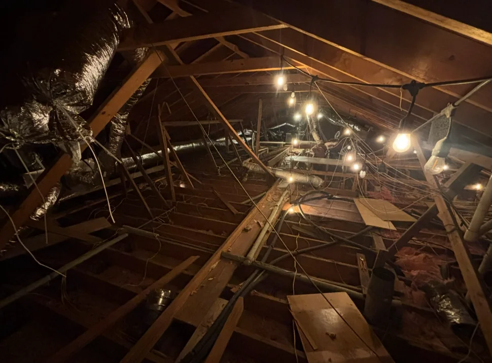 Large unfinished attic with exposed roof framing, ductwork, wiring, and work lights.